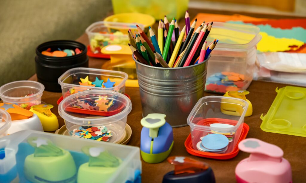 Colored pencils and paper with paint and paper. There are various jugs of paint on a brown crafting table.