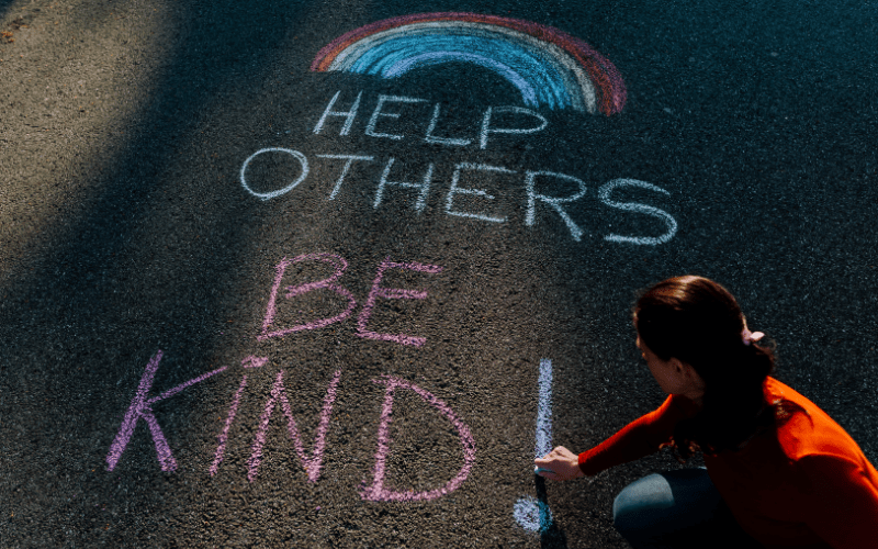 The words help other be kind with a rainbow written by a woman in chalk